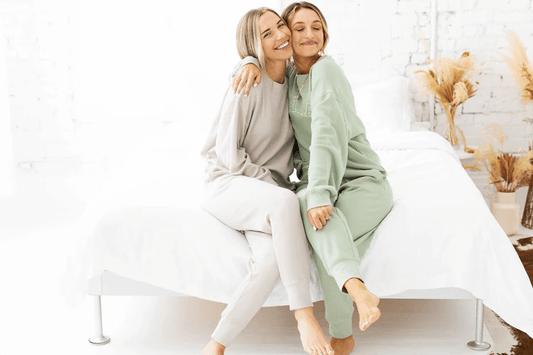 Two women sit on a bed embracing each other in loungewear