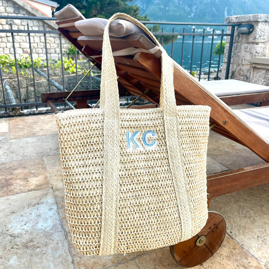 straw tote bag hanging on corner of a wooden lounge chair. tote features a custom embroidery of 2 shadow block initials in a hydrangea blue and powder blue thread
