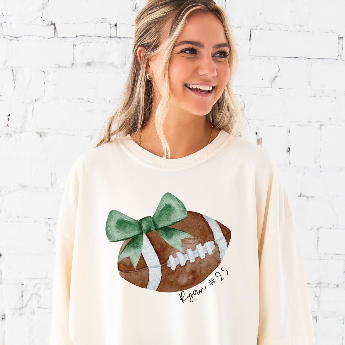 woman wearing an ivory comfort colors t-shirt with a custom watercolor football and green bow print with custom script text along the curve reading Ryan #25