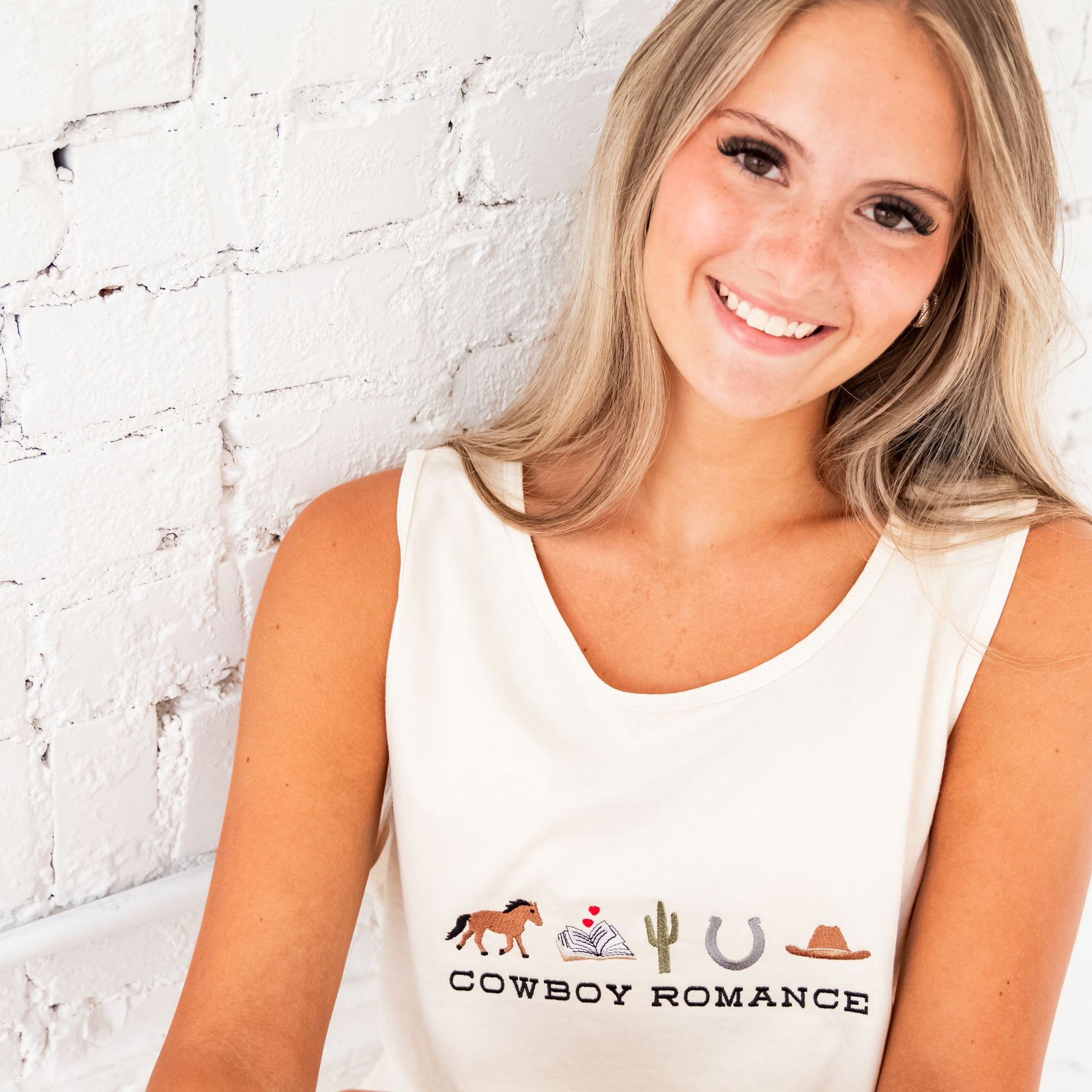 Close up of a blonde woman sitting and leaning against white brick wall wearing an ivory comfort colors tank top with a custom embroidered design on the center chest featuring "cowboy romance" in a western inspired font centered underneath the following western icon: horse, book with a mini heart above, cactus, horseshoe, and cowboy hat.
