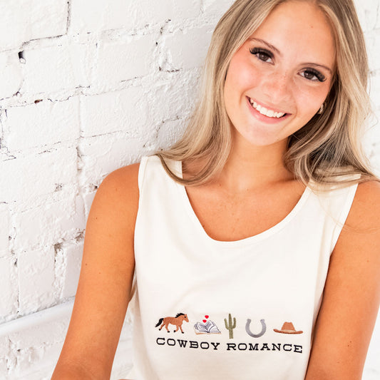 Close up of a blonde woman sitting and leaning against white brick wall wearing an ivory comfort colors tank top with a custom embroidered design on the center chest featuring "cowboy romance" in a western inspired font centered underneath the following western icon: horse, book with a mini heart above, cactus, horseshoe, and cowboy hat.