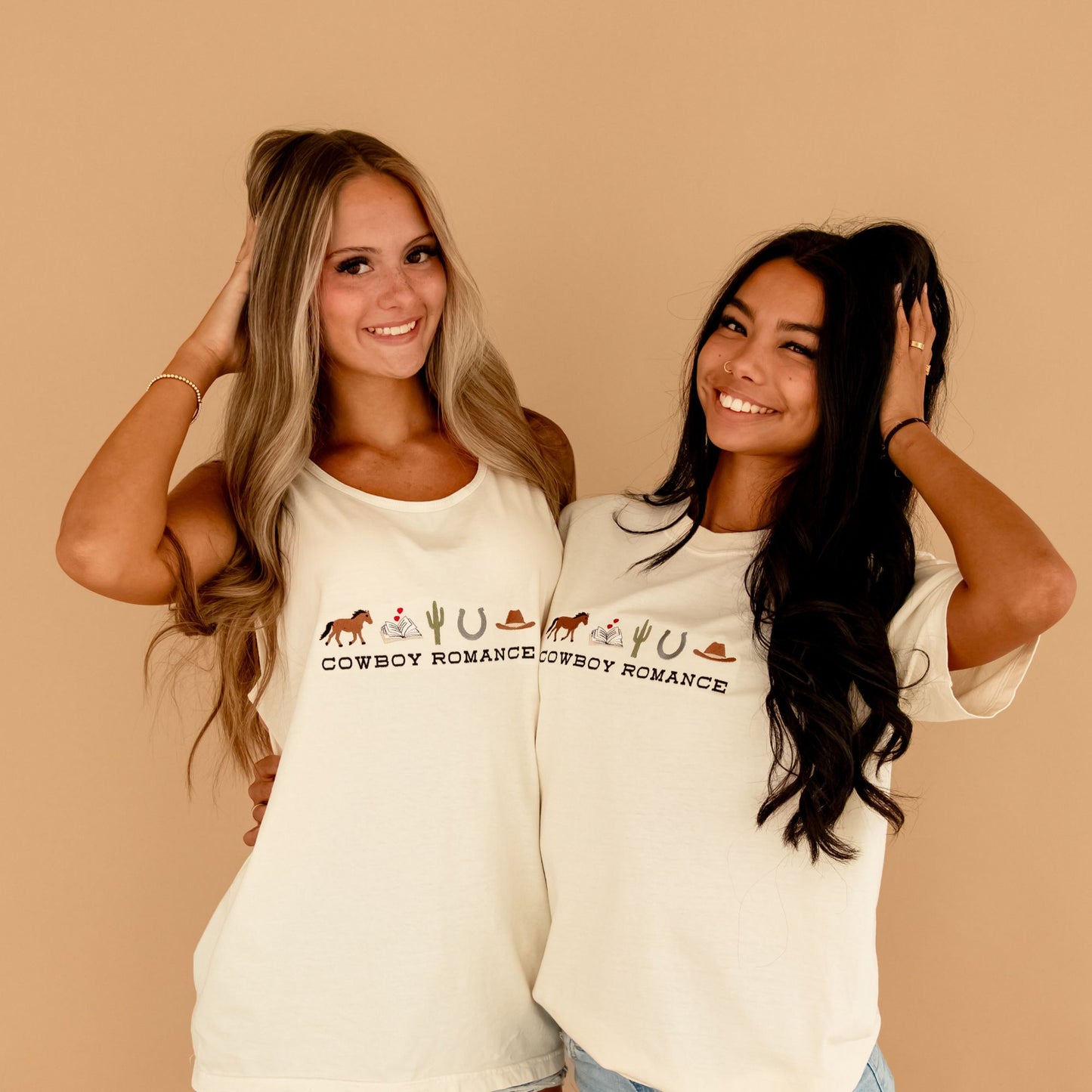 two girls standing side-by-side, one is blonde and the other is a brunette. The blonde is wearing an ivory comfort colors tank top with a custom embroidered design on the center chest featuring "cowboy romance" in a western inspired font centered underneath the following western icon: horse, book with a mini heart above, cactus, horseshoe, and cowboy hat. And the brunette is wearing an ivory comfort colors t-shirt with the same design