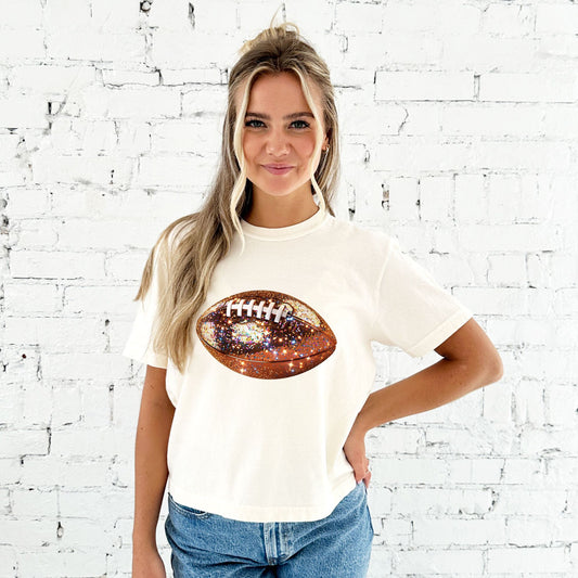 young woman wearing a cropped ivory comfort colors t-shirt with large glitter football design across the chest