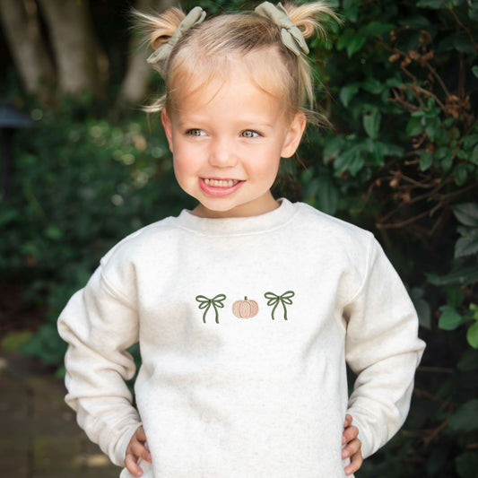 Little girl standing outside wearing a heather oatmeal sweatshirt with bow pumpkin bow embroidered design on the center chest.