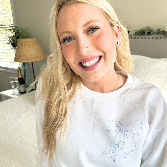 Woman wearing a white sweatshirt with a blue bow design, sitting in a room with a lamp and plant in the background.