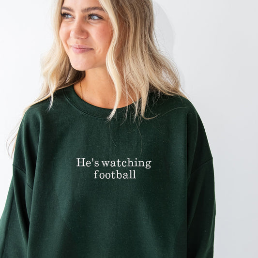 girl wearing a forest green crewneck sweatshirt with custom he's watching football embroidered design on the center chest
