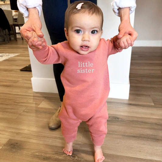 baby wearing a mauve fleece romper with embroidered little sister in white thread on the chest