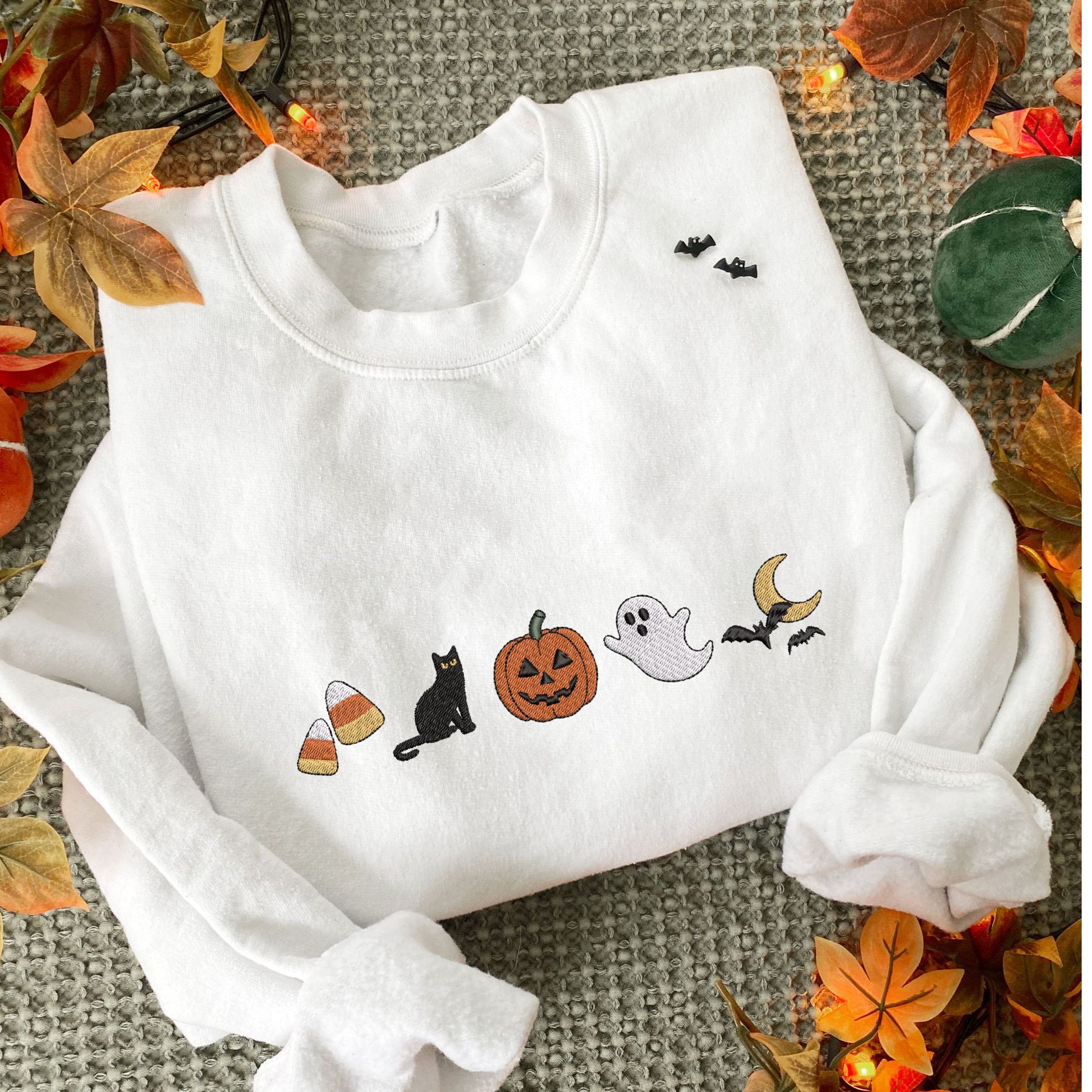 styled fall flat lay photo of a white crewneck sweatshirt with embroidered halloween icons. The icons are embroidered as following: candy corn, black cat, jack-o-lantern, ghost, and bats flying over the moon.