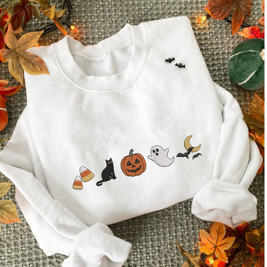 styled fall flat lay photo of a white crewneck sweatshirt with embroidered halloween icons. The icons are embroidered as following: candy corn, black cat, jack-o-lantern, ghost, and bats flying over the moon.