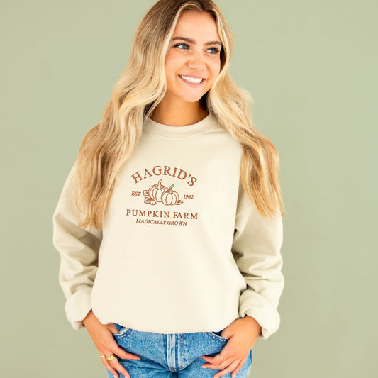 young woman wearing a medium sand crewneck sweatshirt with embroidered hagrid pumpkin farm harry potter inspired design in pumpkin spice thread across the center chest