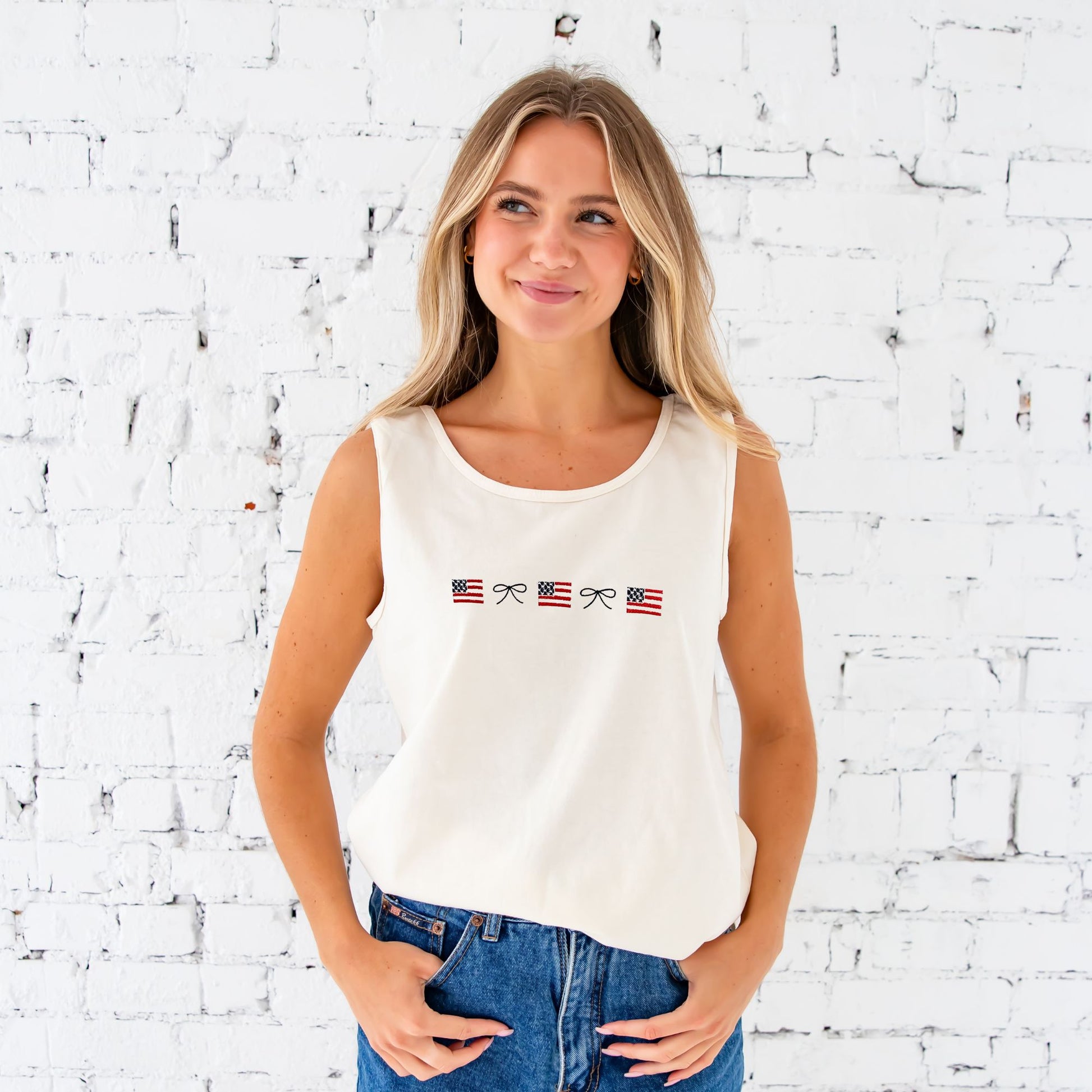 blonde woman standing in front of a white brick wall wearing denim shorts and an ivory comfort colors tank top with embroidered mini usa flags and dainty bow design aross the chest
