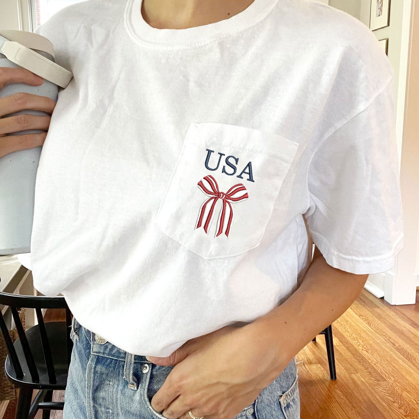 close up of a white comfort colors t-shirt with embroidered red and white striped bow and USA in navy above