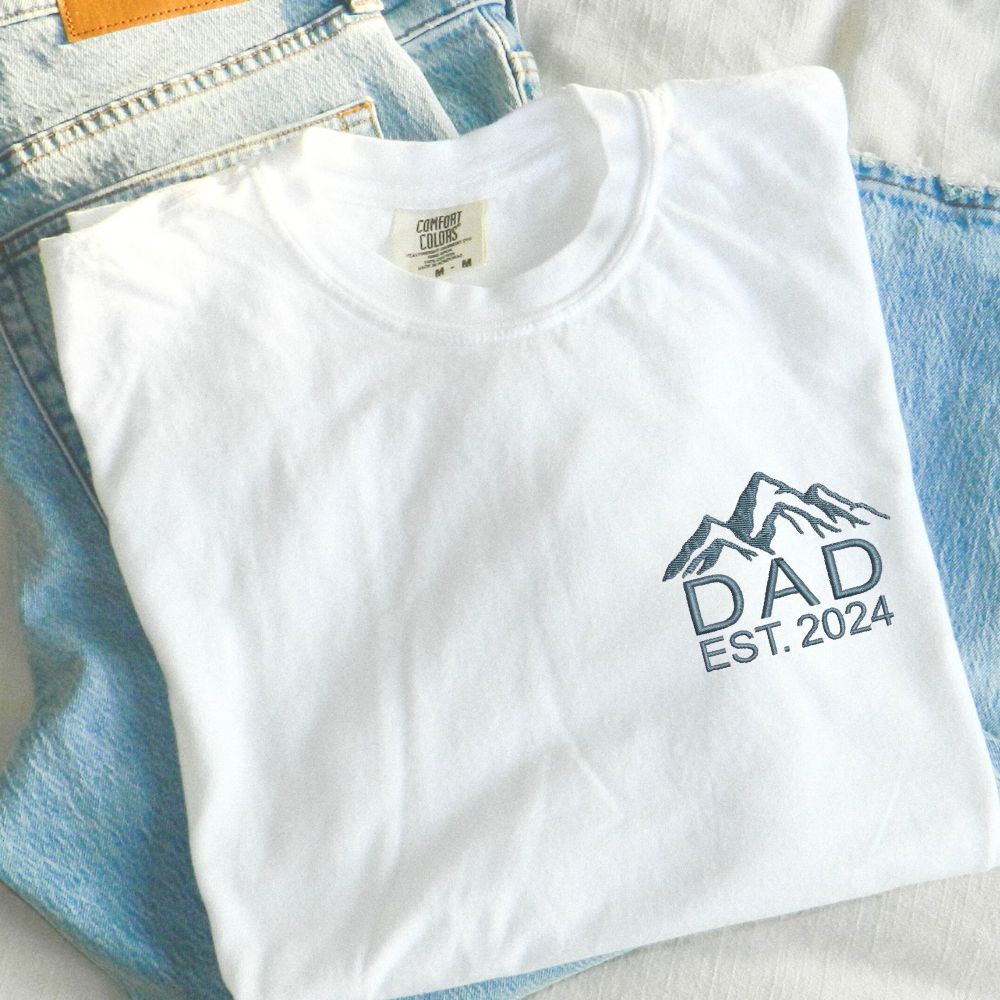 a white t-shirt with custom mountain icon and dad/custom year embroidered design on the left chest in French blue thread