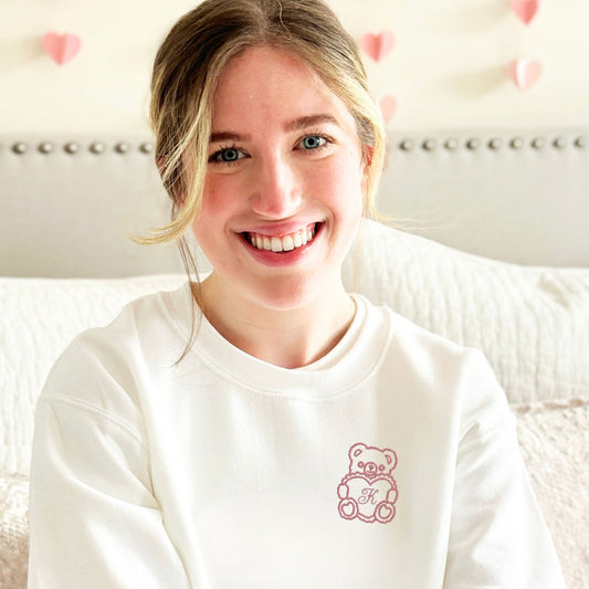girl wearing a white crewneck sweatshirt with a design of an outlined teddy bear holding a heart with an initial in the center embroidered on the left chest in mauve thread