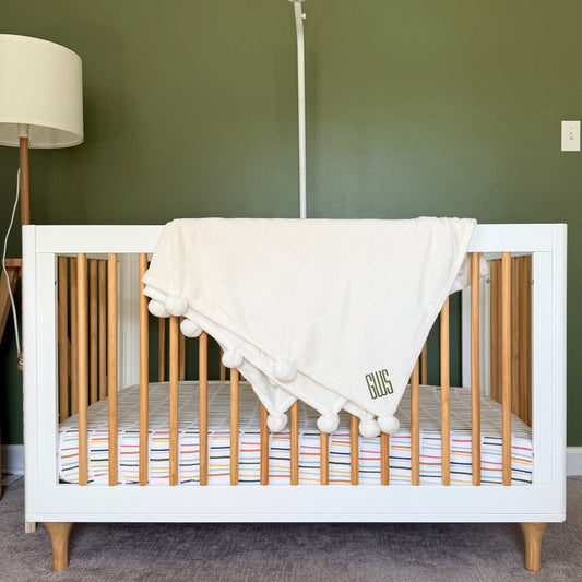 soft white pom pom blanket draped over the edge of a crib with custom monogram embroidered design