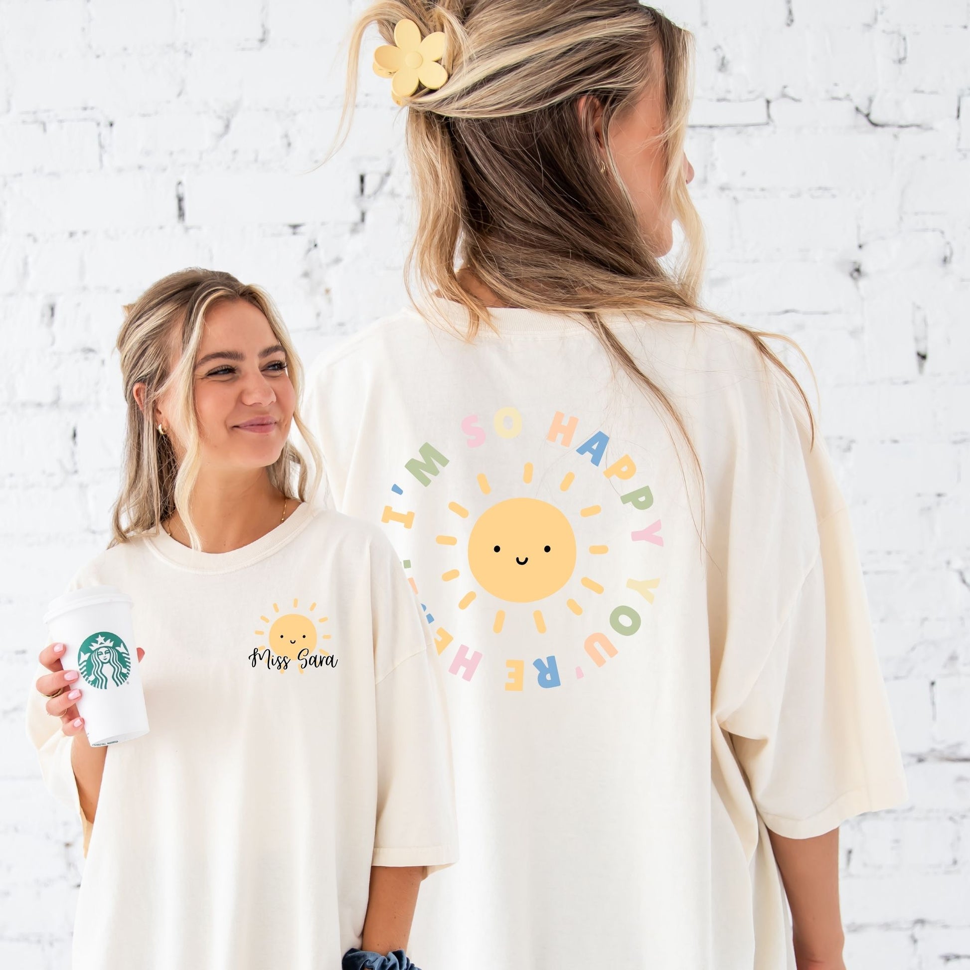 Girl wearing an oversized ivory comfort colors t-shirt. the front features a left chest print with a cute smiling yellow sun and a custom script name in black ink. on the back is a large round print with text in multiple colors saying "I'm glad you're here" surrounding a smiling yellow sun