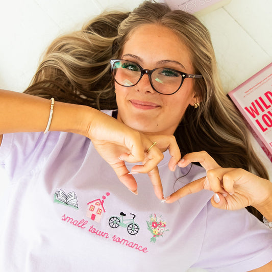 girl laying on her back on the ground with books surrounding her and she's wearing an orchid comfort colors t-shirt with a cute small town romance embroidered icon design on the center chest featuring: an open book, a house with hearts coming out of the chimney, a vintage bike, and a bouquet of colorful flowers. She's smiling, wearing glasses, and holding her hands/fingers up in a heart shaped pose.