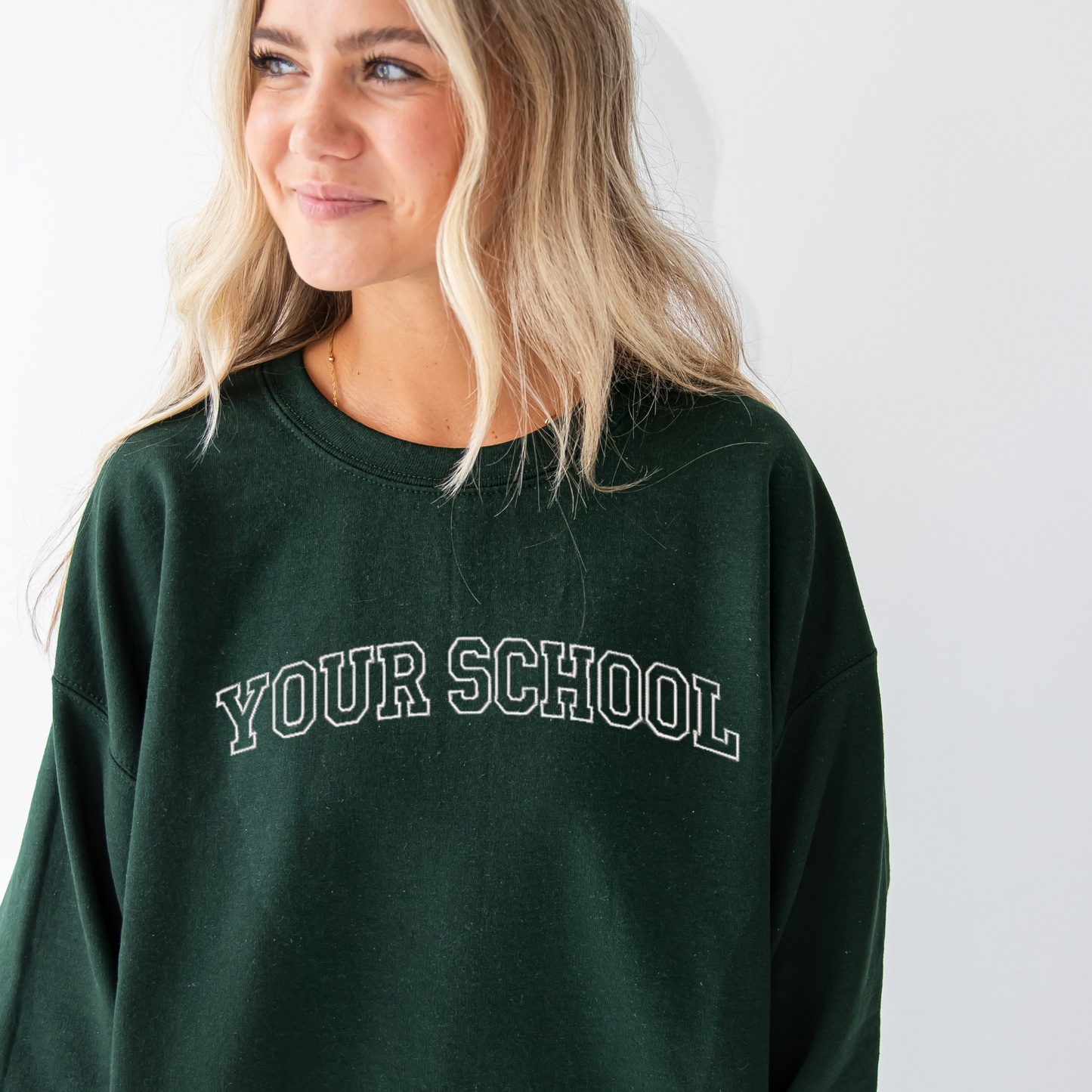young woman wearing a forest green crewneck sweatshirt with custom athletic block outline embroidered font across the chest