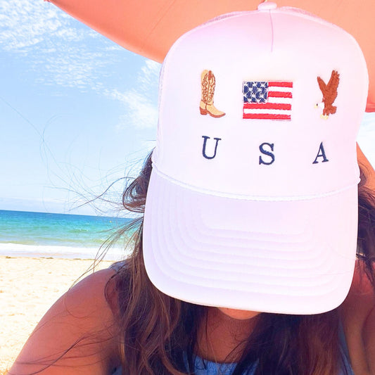 woman on the beach under an umbrella wearing a white trucker hat with custom cowboy boot, usa flag, and bald eagle USA embroidered design