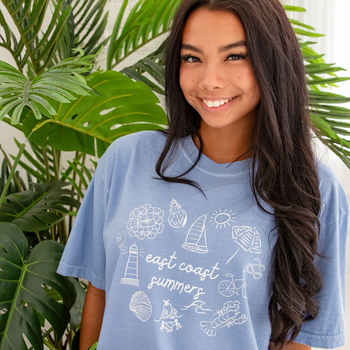 Woman standing wearing washed denim comfort colors shirt with a custom design featuring a flower, shell, sailboat, sun, umbrella, lighthouse, clam, swimsuit, waves, lobster, bicycle, sunglasses, and east coast summer printed at the center in a script font.
