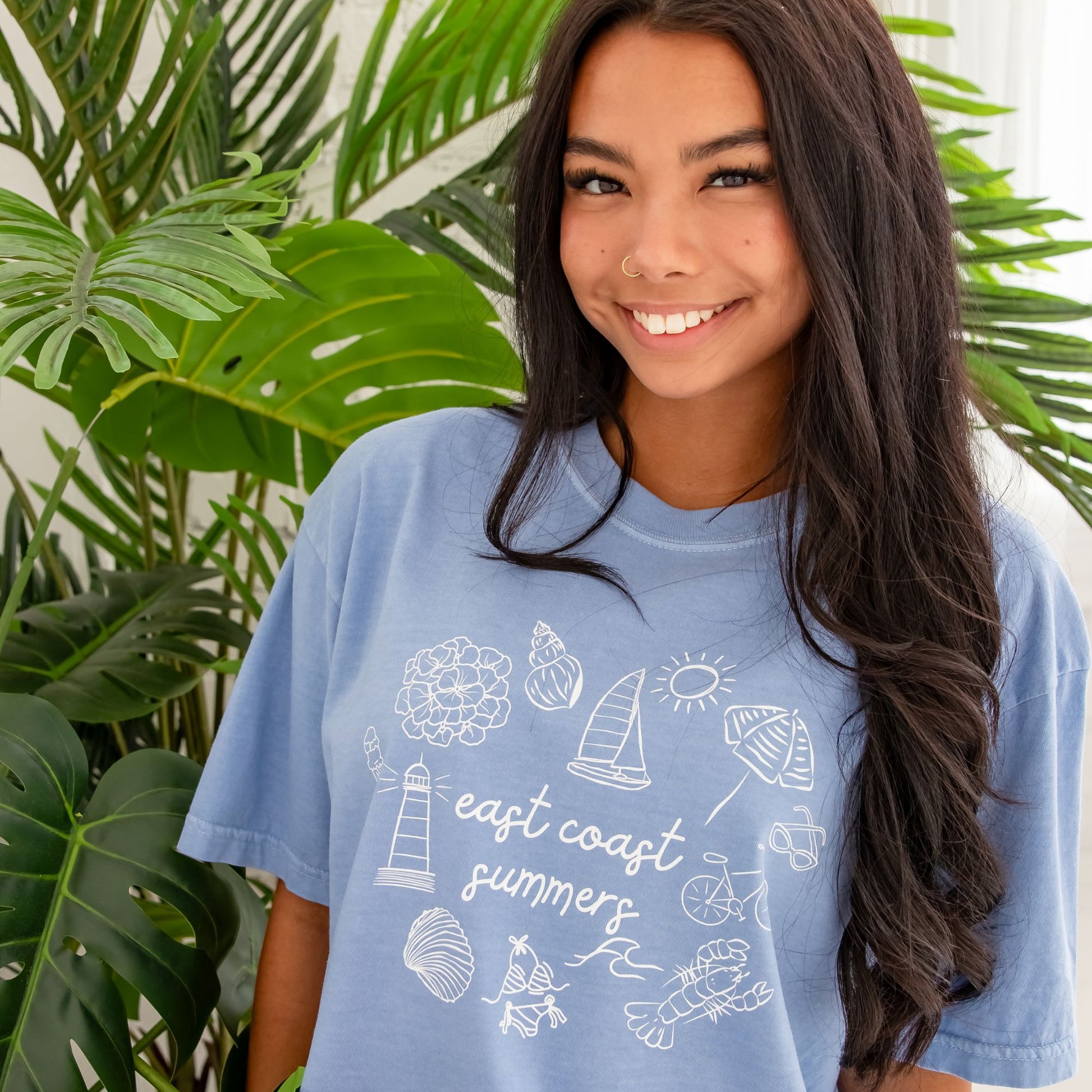 Woman standing wearing washed denim comfort colors shirt with a custom design featuring a flower, shell, sailboat, sun, umbrella, lighthouse, clam, swimsuit, waves, lobster, bicycle, sunglasses, and east coast summer printed at the center in a script font.