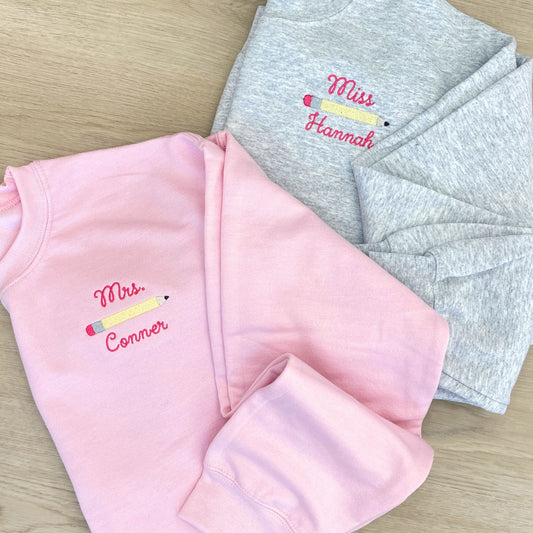 Pink and gray sweatshirts with embroidered names and pencil design on the left chest laying wooden floors