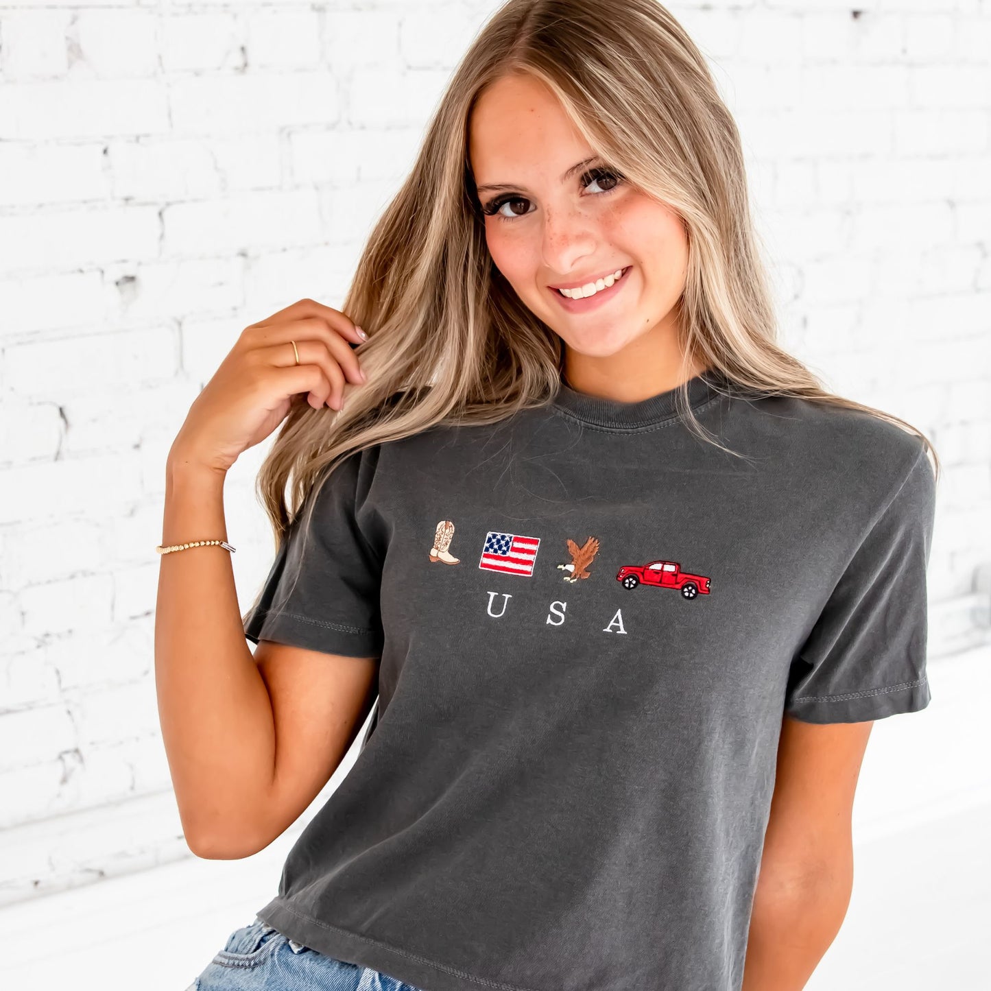 A girl with long blonde hair wearing a cropped pepper comfort colors tee featuring mini cowboy boot, american flag, bald eagle, and pickup truck embroidered icons and minimal USA text embroidered in white under the icons. She's wearing denim shorts and sitting on a white stool in front of a white brick wall.