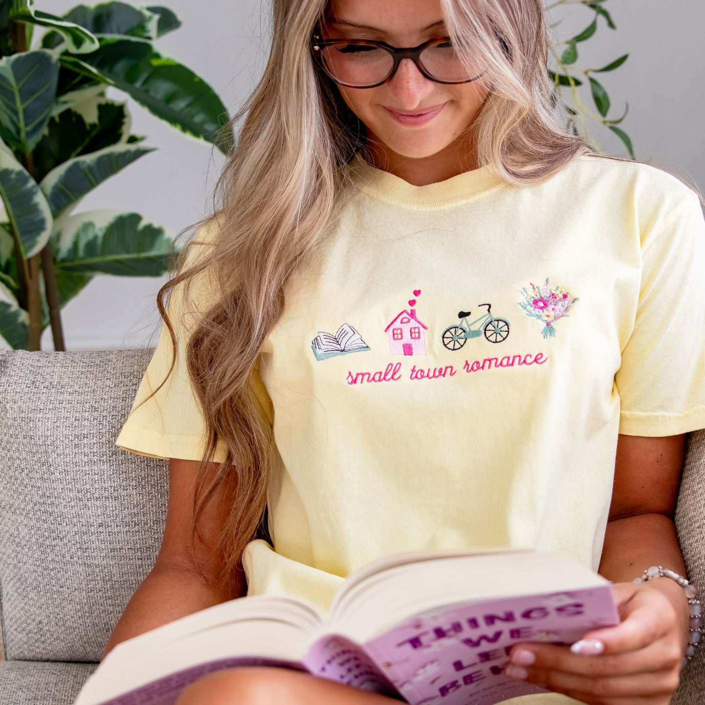 girl with long blonde hair sitting on a couch reading and wearing glasses and a banana comfort colors t-shirt with a custom small town romance design embroidered on the center front featuring an open book, house with hearts coming out of the chimney, bike, and flower bouquet icons.
