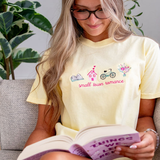 girl with long blonde hair sitting on a couch reading and wearing glasses and a banana comfort colors t-shirt with a custom small town romance design embroidered on the center front featuring an open book, house with hearts coming out of the chimney, bike, and flower bouquet icons.