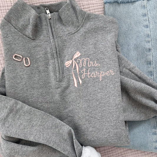 oxford gray quarter zip sweatshirt folded and laying on a pink muslin blanket paired with gold hoops and light blue jeans. quarter zip features a long tail bow and curly script name embroidered design on the left chest in powder pink thread.