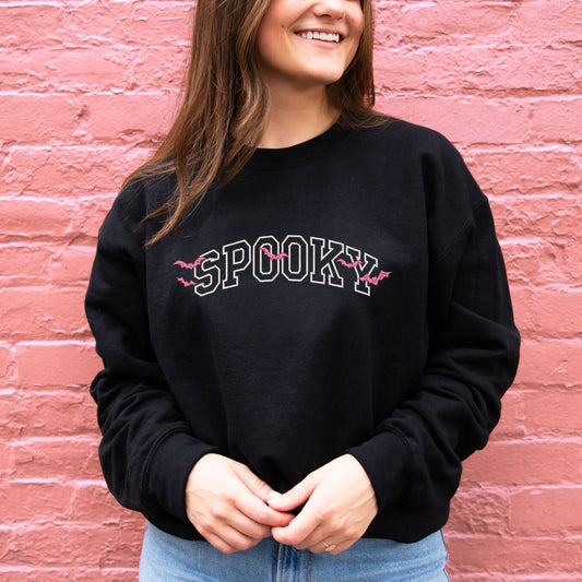 young woman wearing a black crewneck sweatshirt with embroidered SPOOKY in white thread and bats in pink thread overtop the design