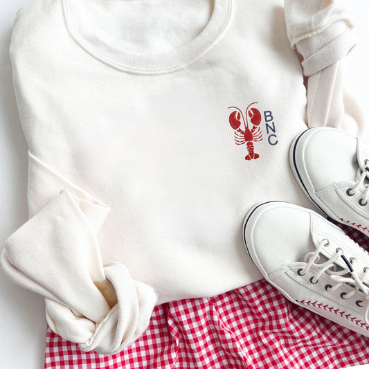 Flat lay of sweet cream sweatshirt with lobster icon embroidered at the left chest with custom monogram design embroidered at the left of the lobster. Partnered with white sneakers and gingham red and white shorts.