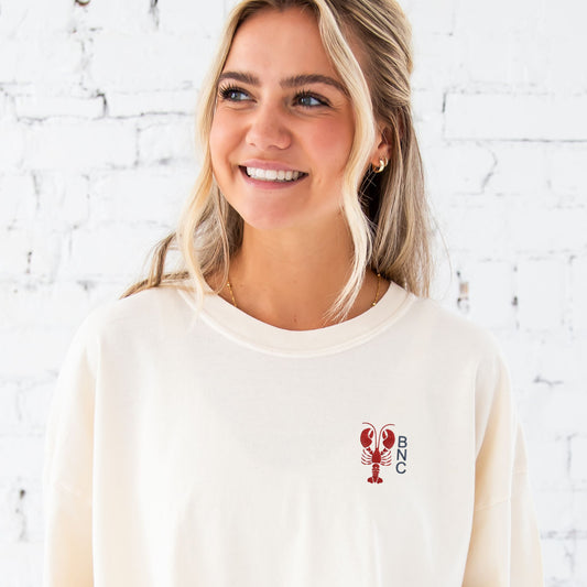 Girl standing in front of a white brick wall wearing an oversized ivory comfort colors t-shirt with a custom embroidered design on the left chest featuring a mini lobster and 3 initials