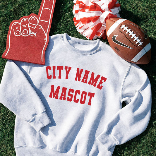 White sweatshirt with 'CITY NAME MASCOT' text, foam finger, pom-pom, and football on grass.