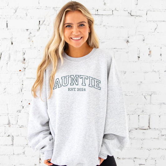 young woman wearing an ash crewneck sweatshirt with embroidered AUNTIE in all caps athletic block font across the chest  and Est. 2024 underneath in  french blue  thread.