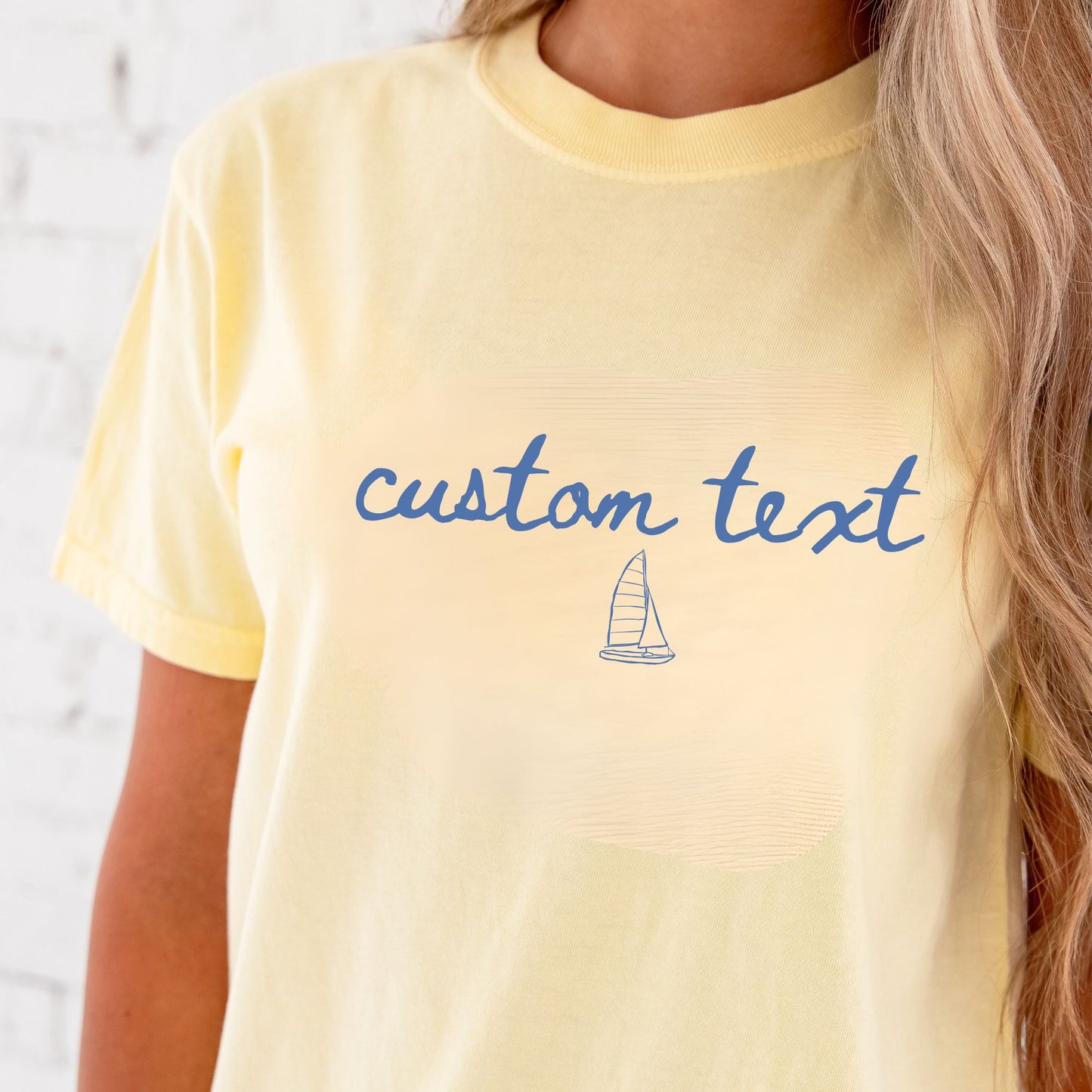 close up of woman wearing a butter short sleeve comfort colors t-shirt with personalized custom text and sailboat printed design on the center chest in a soft blue ink.