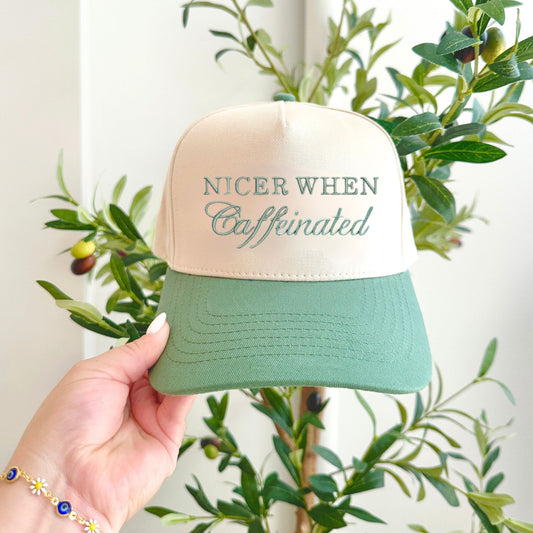 woman holding up a sage green two tone canvas trucker hat in front of a eucalyptus tree. Hat features a mixed text font embroidered design reading nicer when caffeinated