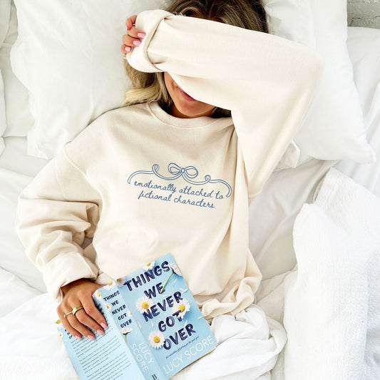 girl laying in bed reading and wearing a sweet cream crewneck with 'emotionally attached to fictional characters' bow design printed across the chest in french blue ink