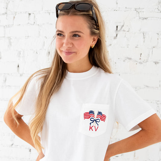 Woman standing in front of a white brick wall. She's wearing a white comfort colors pocket tee with a cute crossed American flags and bow monogram embroidered design on the pocket. Thread colors are a classic red and navy with a touch of camel for the flag poles. She's wearing her hair half up, and sunglasses are propped on the top of her head.