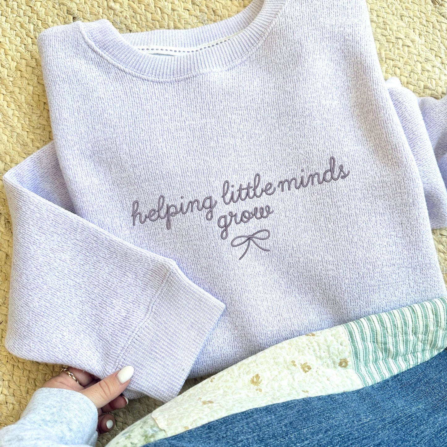 ice purple cozy knit nantucket fleece sweatshirt folded and laying on a jute woven rug. The sweatshirt features a cute embroidered helping little minds grow design with a small delicate bow underneath. Embroidery is in smoky orchid thread.