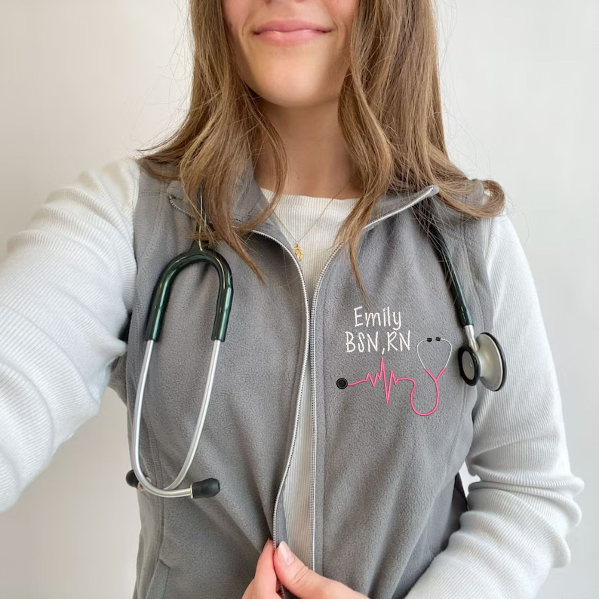young woman wearing a light weight grey fleece vest with embroidered nurse heartbeat stethoscope in pink thread  on the left chest 
 with name and credentials in white thread.