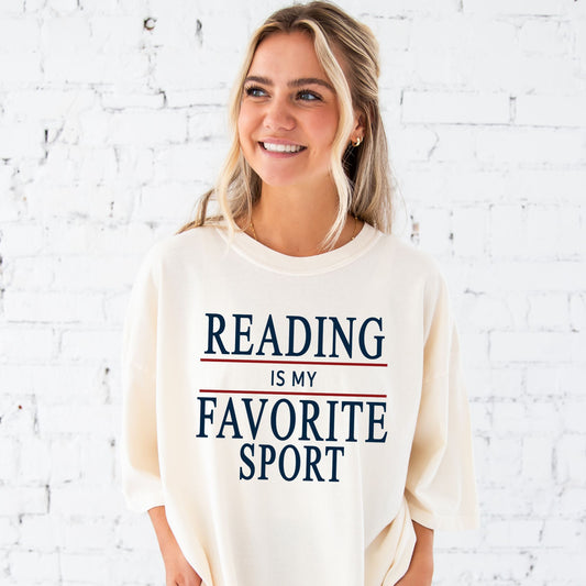 woman standing in front of brick wall wearing ivory shirt with a reading is my favorite sport printed design in navy and red ink
