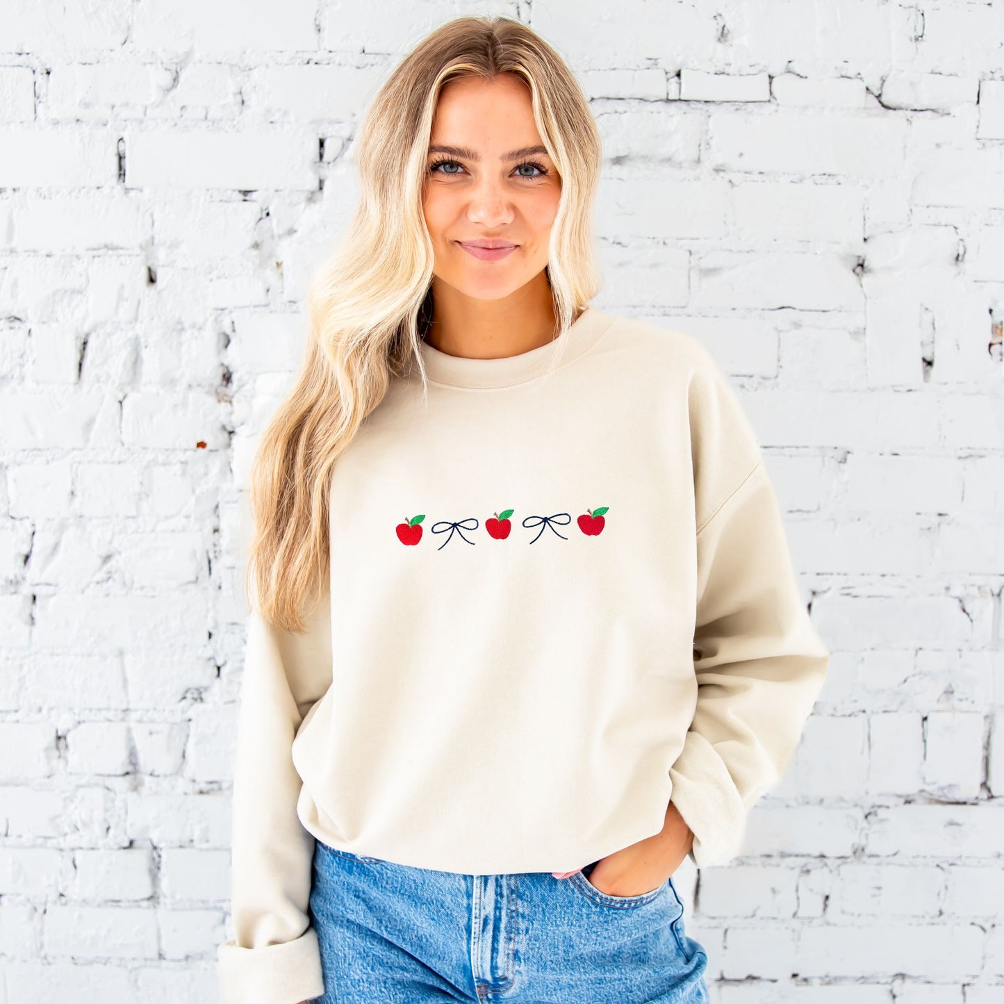 Blonde woman standing in front of a white brick wall wearing jeans and a sand crewneck sweatshirt embroidered with navy dainty bows and red apples in an alternating pattern.