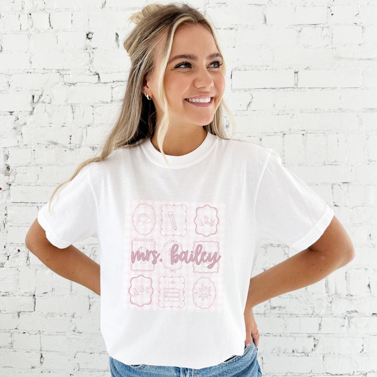 woman standing in front of a white brick wall wearing a white comfort colors t-shirt with custom print. print features a pale pink gingham pattern background with nine bow frames with cute line sketch teacher icons. Also features custom script text name across the center of the design in a darker pink ink.