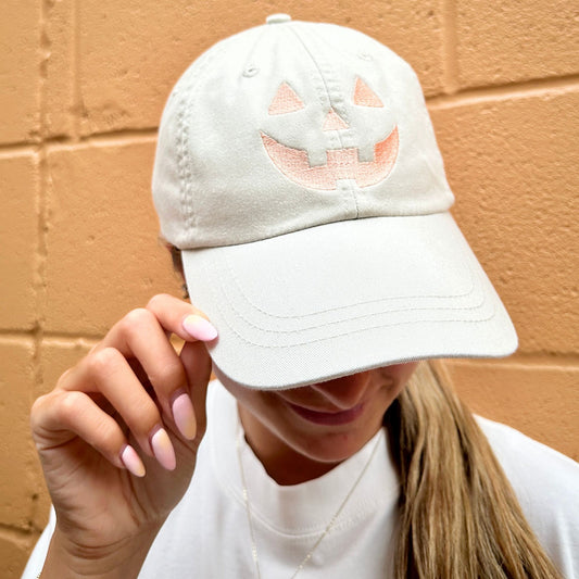 Person wearing an ivory pigment-dyed cap with a pumpkin face embroidered design, and she's standing against an orange brick wall.
