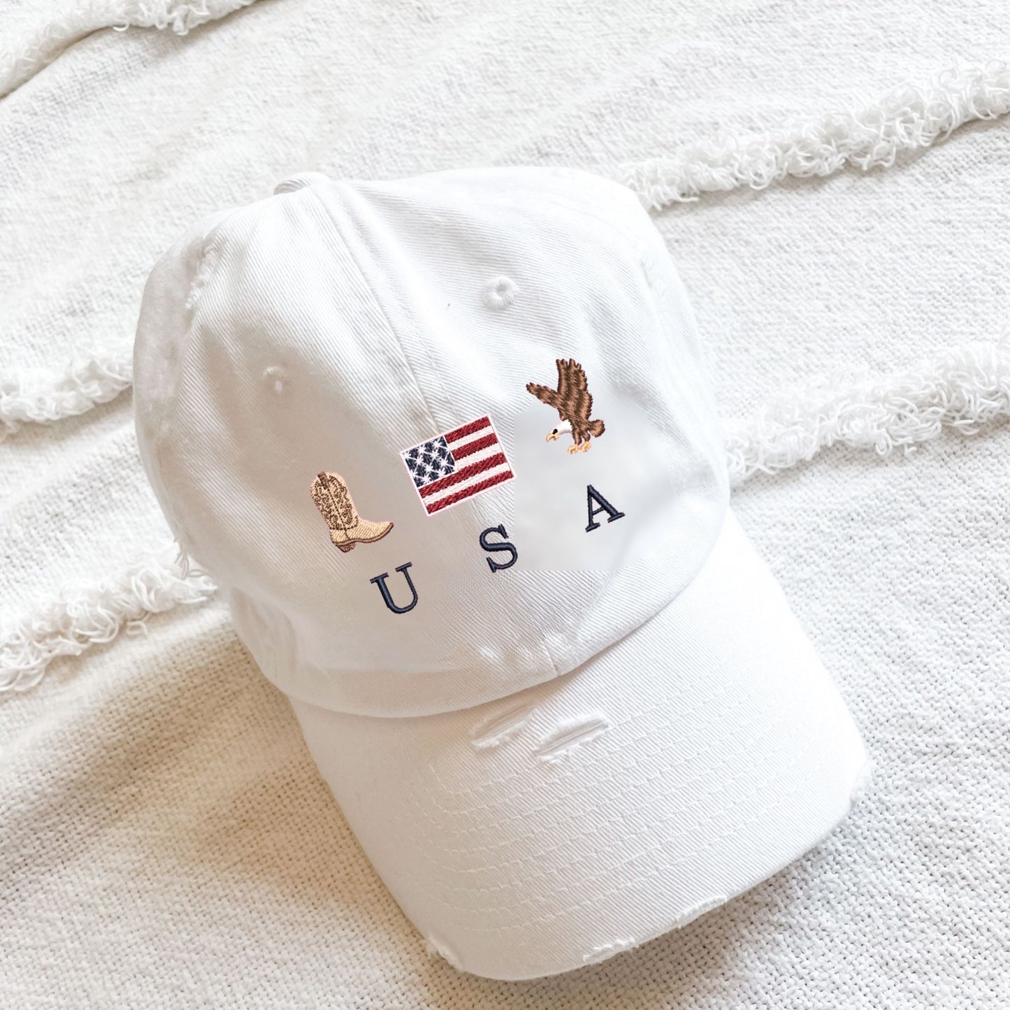 a white distressed baseball cap with embroidered cowboy boot, american flag, eagle, and USA design
