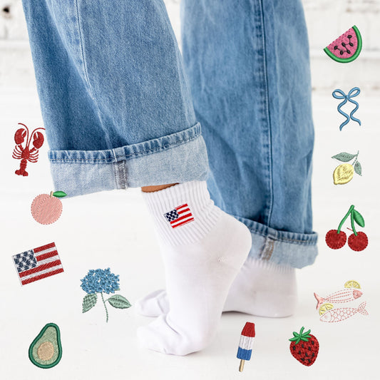 person wearing blue jeans rolled up to show off white crew socks featuring a mini usa flag embroidered icon. Person's legs are surrounded by lobster, peach, hydrangea, avocado, popsicle, strawberry, sardines, cherries, lemon, ribbon bow, watermelon embroidered icons.