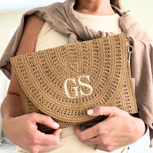 woman wearing a tank top with a sweater over her shoulders holding a woven taupe straw fold-over clutch purse featuring a two letter embroidered monogram in natural thread