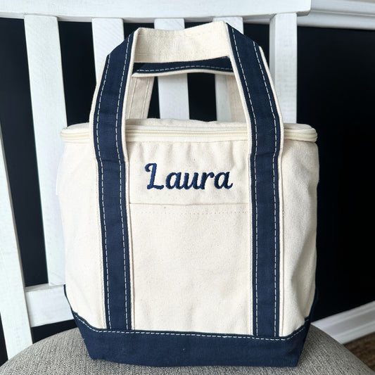 Beige tote bag with navy handles and trim, embroidered with 'Laura', on a white bench.