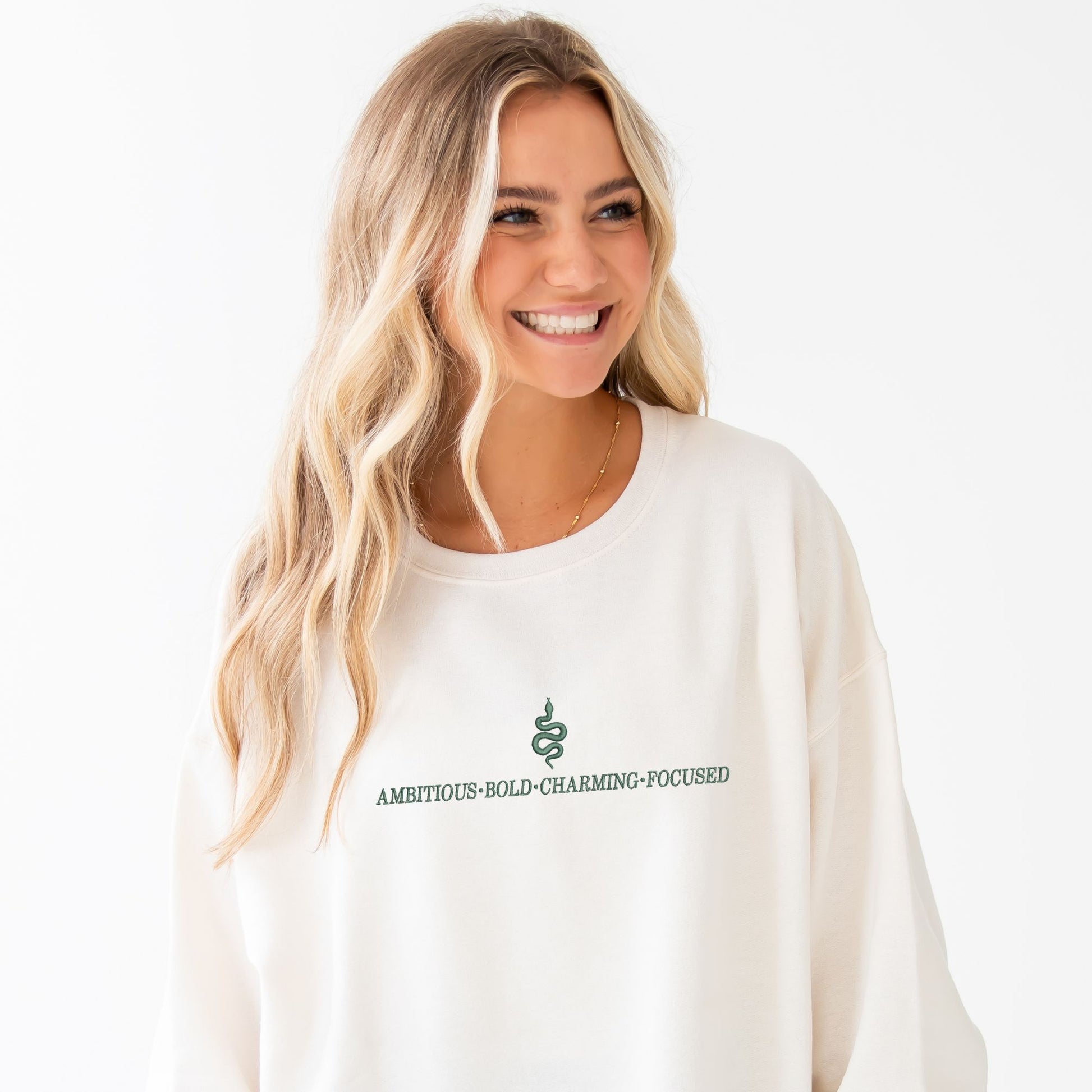 Girl wearing a sweet cream crewneck sweatshirt with custom embroidered Slytherin snake and text reading ambitious, bold, charming, focused.